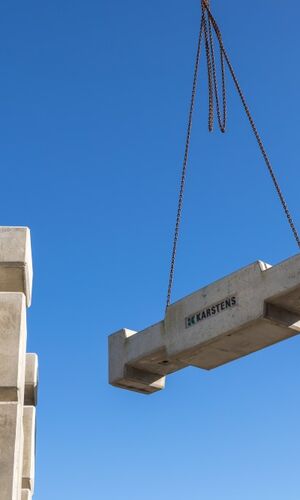 Betonfertigteil mit der Aufschrift 'HK Karstens' wird von einem Kran gehoben, Stapel weiterer Betonfertigteile im Hintergrund