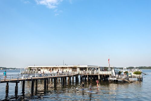 Steg mit Menschen und einem Gebäude am Ende, darüber blauer Himmel, darunter Wasser mit drei Paddlern