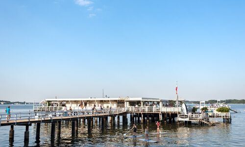 Steg mit Menschen und einem Gebäude am Ende, darüber blauer Himmel, darunter Wasser mit drei Paddlern